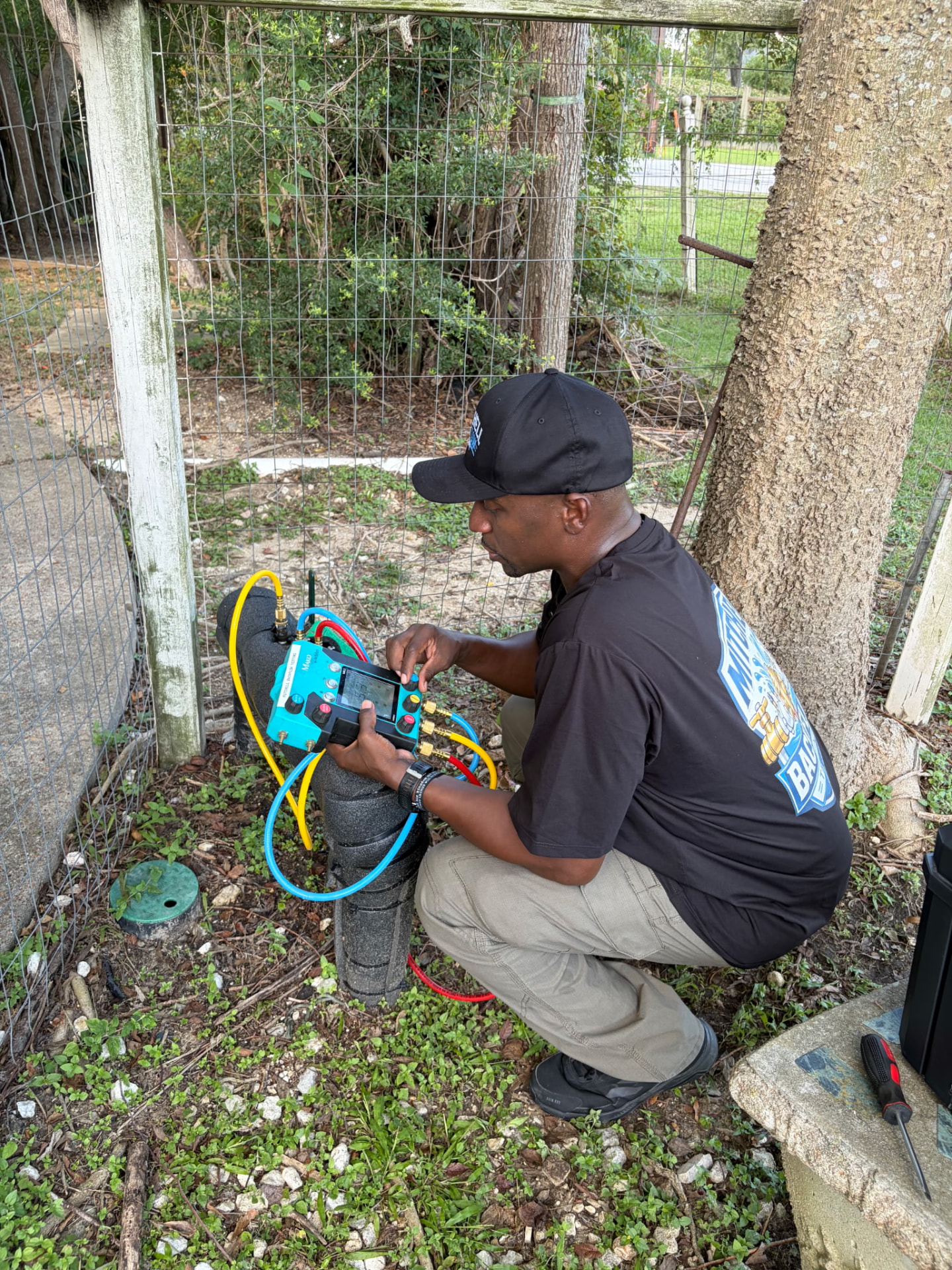 Mitchell Backflow Testing technician crouched beside an insulated backflow assembly, testing it with a digital gauge kit and color-coded hoses
