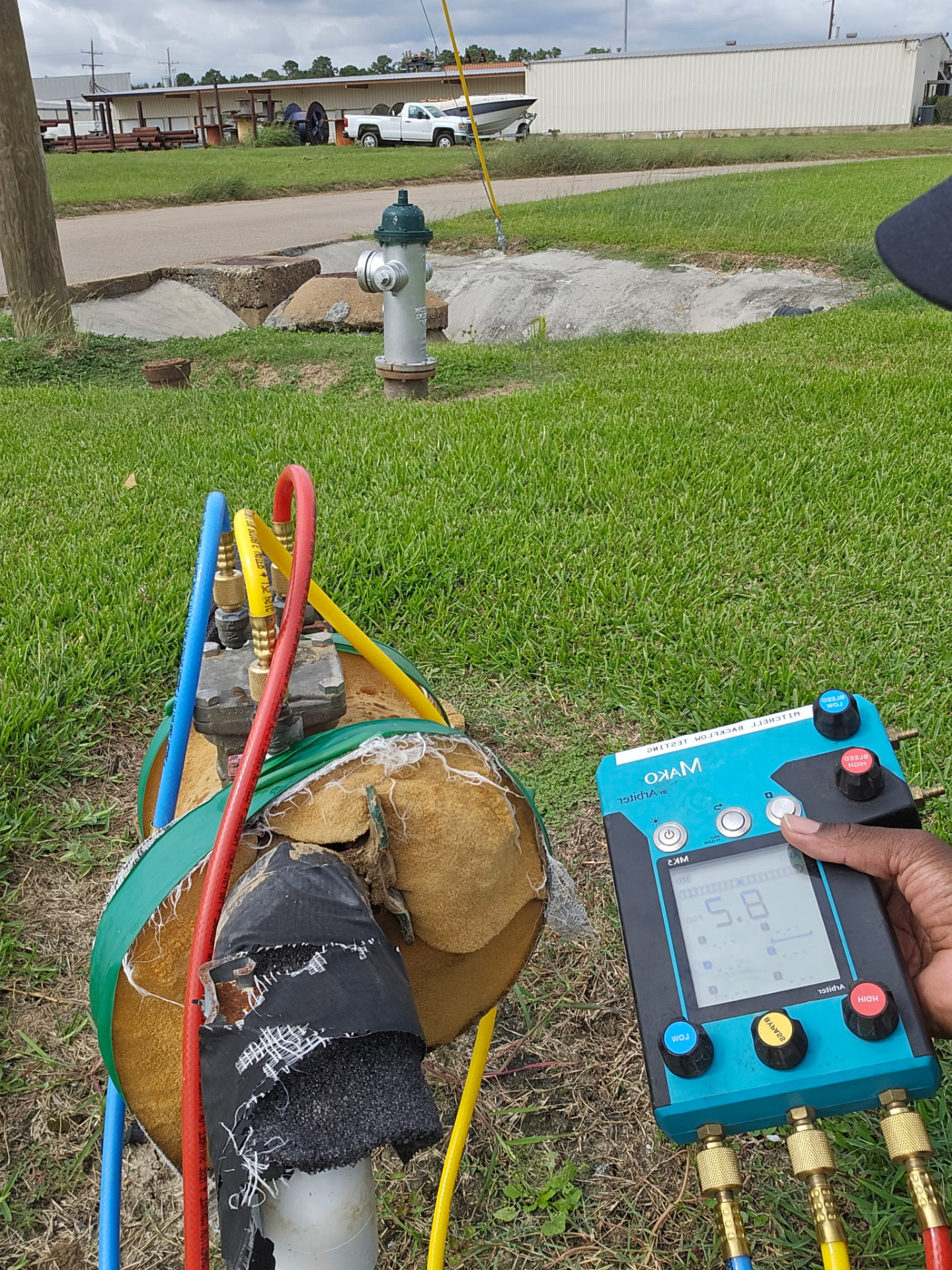 Digital backflow test gauge held on site with hoses running to an insulated assembly, roadway behind