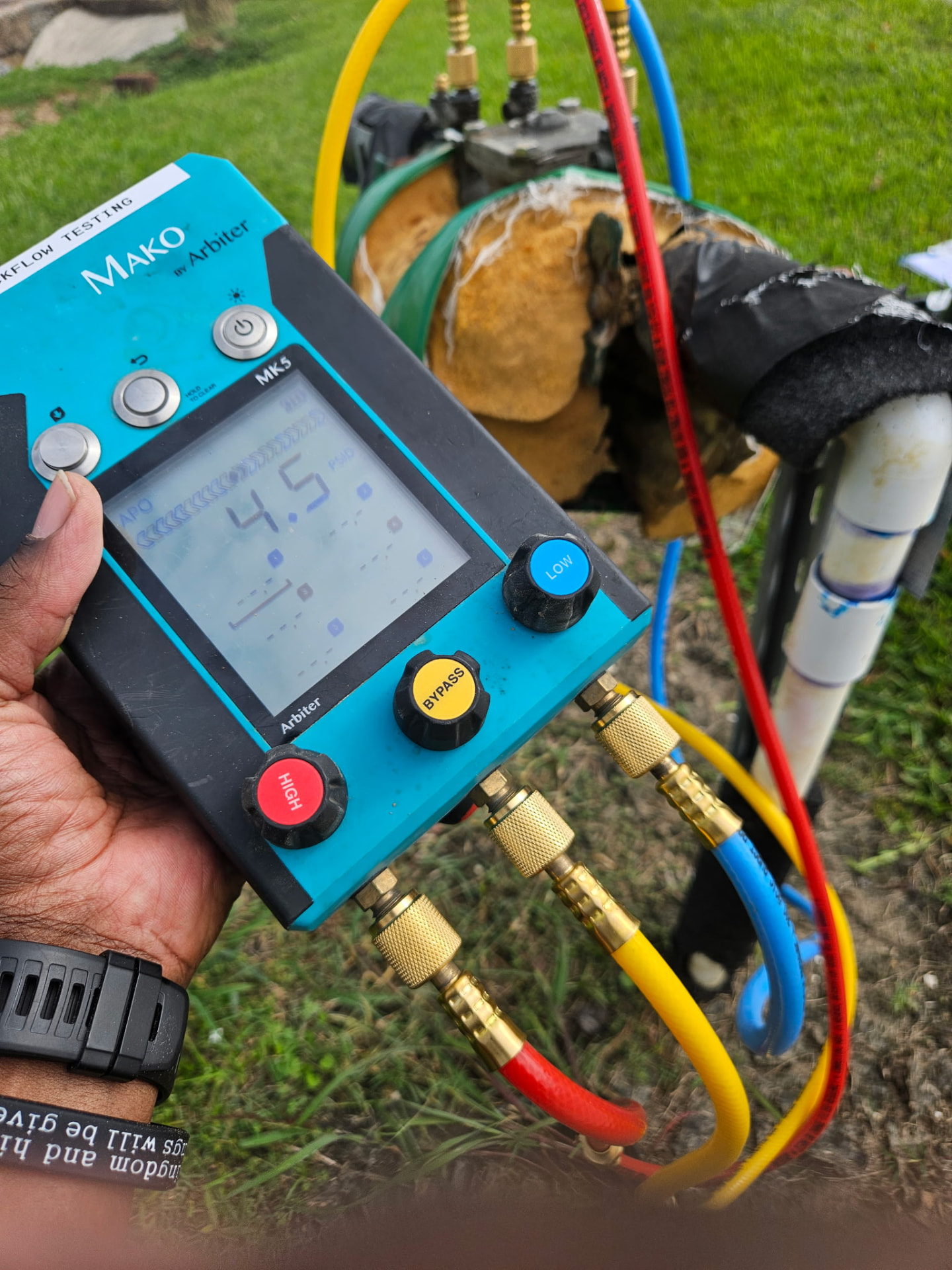 Close view of a digital test gauge connected by red, yellow and blue hoses to an above-ground backflow preventer