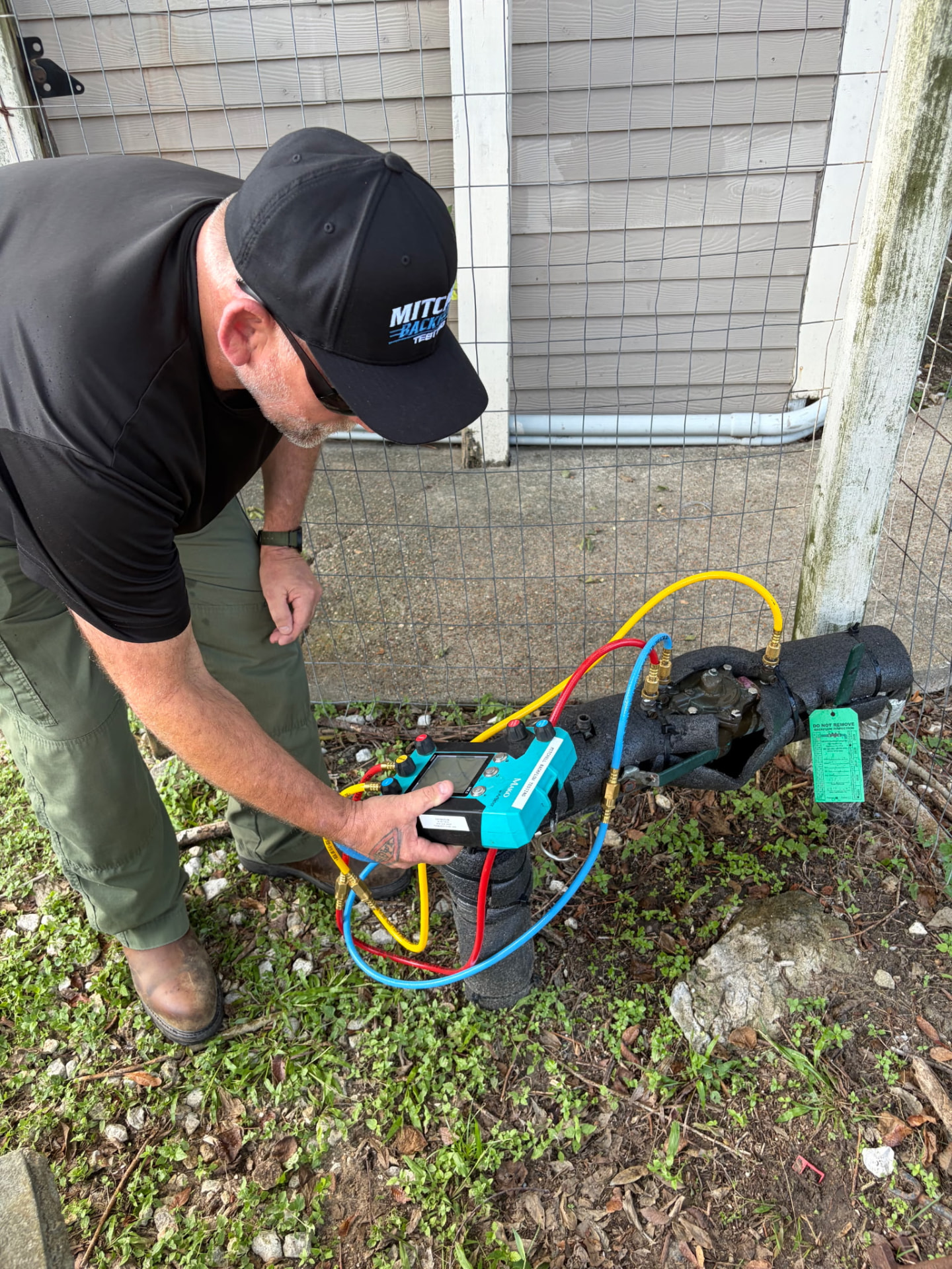 Mitchell Backflow Testing technician bent over a backflow preventer along a wire-fenced property line, reading the digital test gauge
